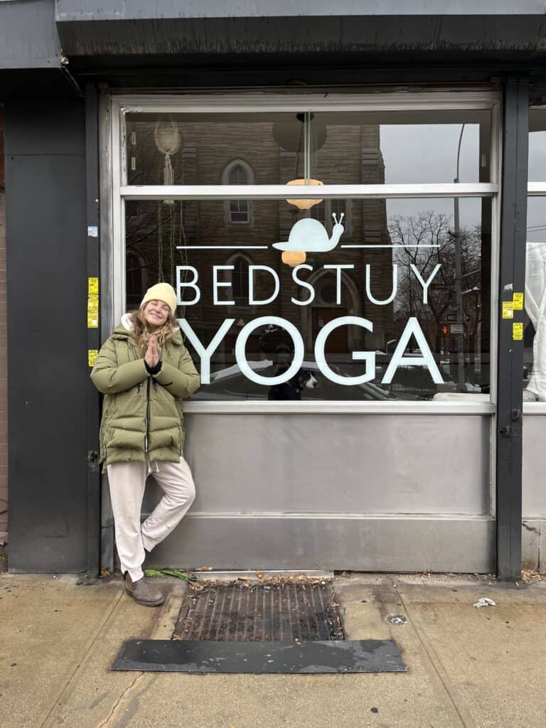 Yoga studio front entrance with a smiling woman in winter clothing, promoting Bedstuy Yoga in Brooklyn.