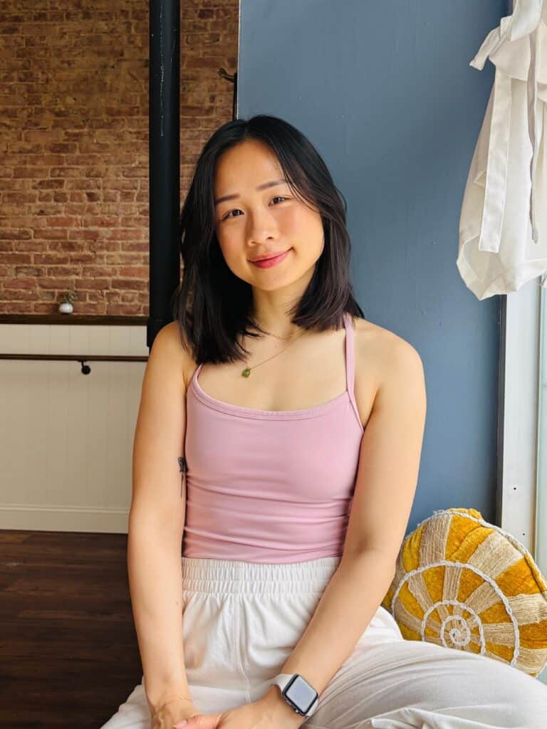 Yoga woman in pink top and white pants, smiling in studio setting.